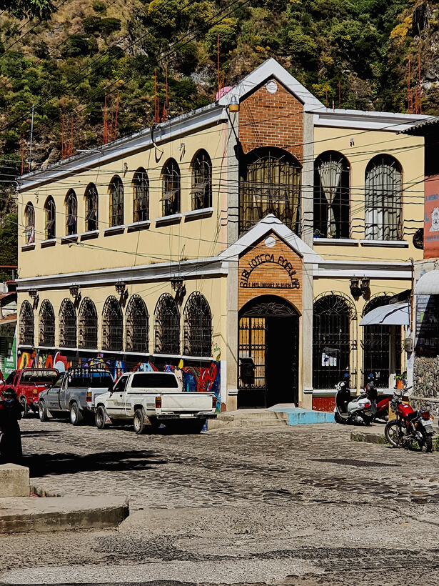 Picture of the Biblioteca Popular in Panajachel. The building is light stone with many arched windows along the side and an arched entrance with a pointed roof. Above the door it reads "Biblioteca Panajachel" on a metal sign. Cars line the street.