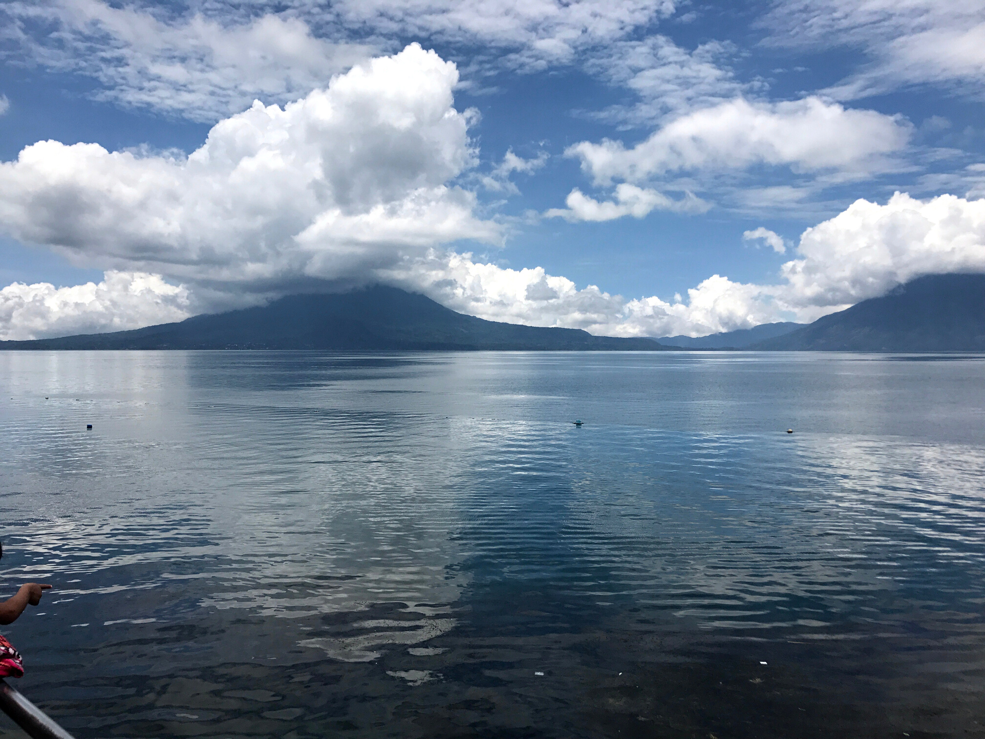 A picture of the waters of beautiful Lake Atitlán reflecting the puffy, white clouds above. Volcán Toliman is in the background as well as Volcán San Pedro.
